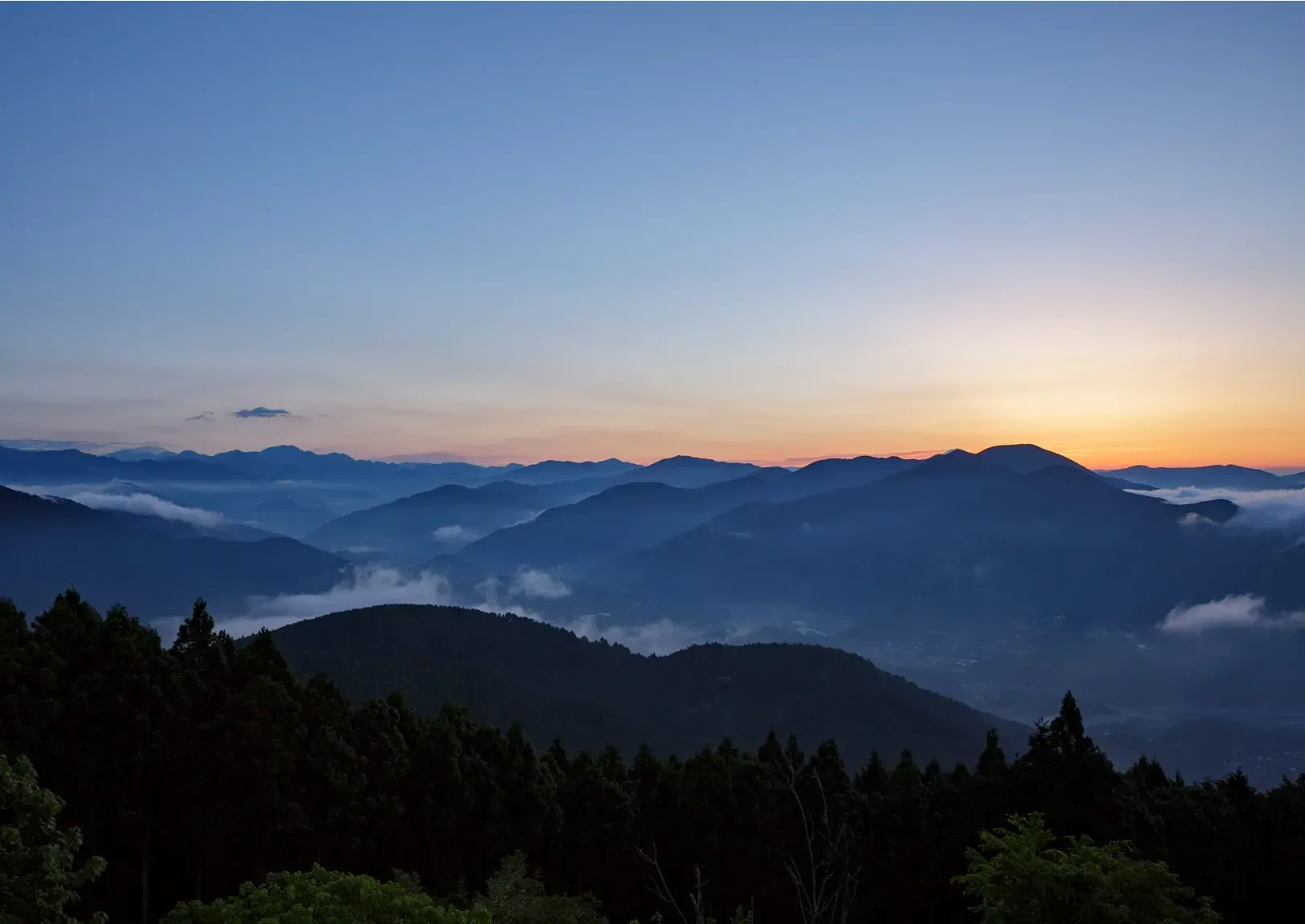 Dawn breaking over layered mountain ridges, cloud lying in the valleys below a dark cedar forest