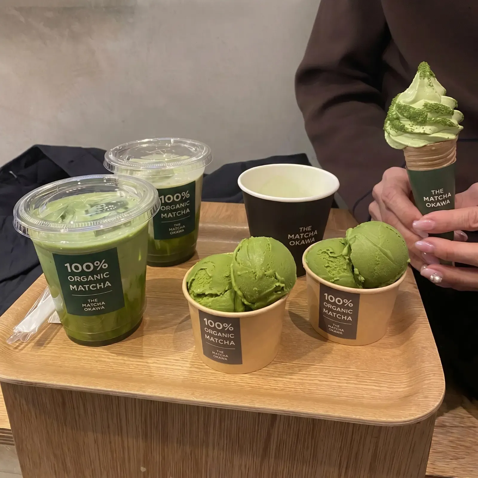 A wooden tray of iced matcha drinks, tubs of green soft-serve and paper cups of ice cream, with a soft-serve cone held above it