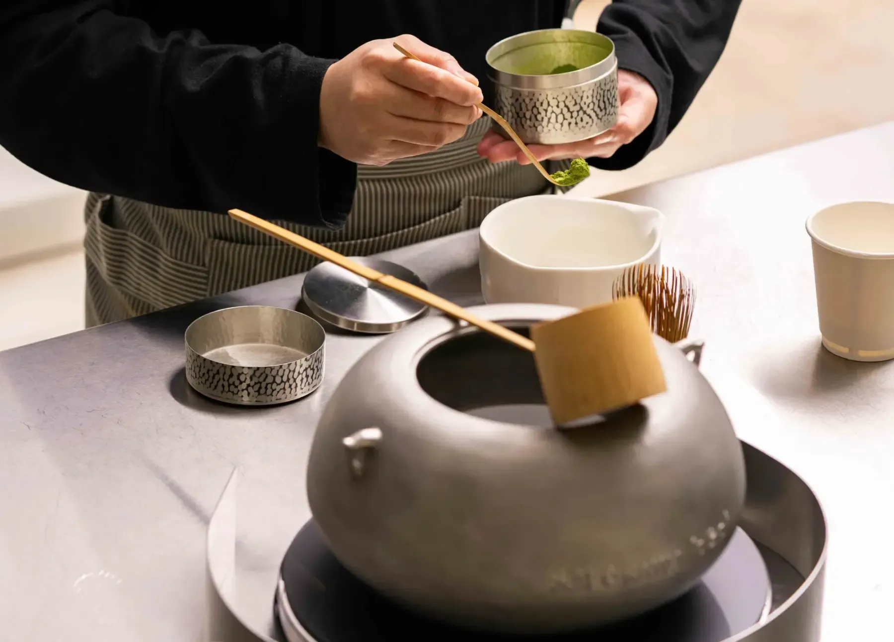 Hands in a dark apron scooping matcha from a hammered tin with a bamboo scoop, above an iron kettle with a bamboo ladle, a whisk and a white bowl on the counter
